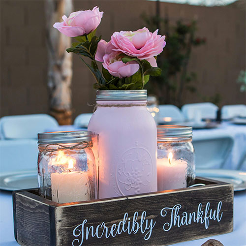 Centerpiece made of 2 candles in glass jars with large pick jar with pink carnations set in a rustic would tray that reads incredibly thankful.