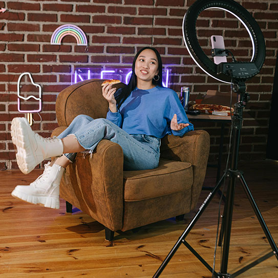 Woman sitting sideways in an armchair with decorative neon signs behind her filming herself for a video blog using a ring light.